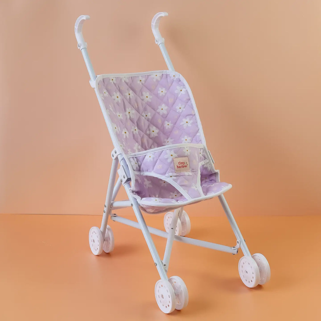 A miniature toy baby stroller with a light purple quilted fabric seat decorated with a white daisy pattern, set against a solid peach background.