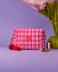 The Somewhere Co Red and white checkered cosmetic bag with a pink flower and lipsticks on a purple background