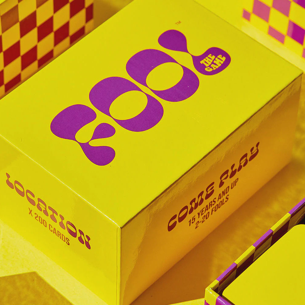 Bright yellow and purple packaging for a card game, with a large box labeled 'come play' and 'LOCATION' in bold purple lettering. The scene is styled with matching yellow and purple checkerboard cards and boxes around it.