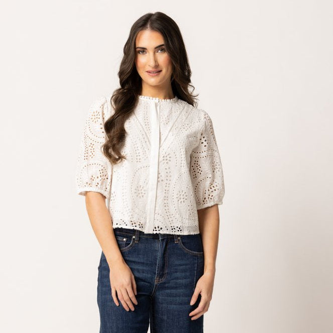 A woman modeling a white short-sleeve crochet knit top with dark blue wide-leg jeans and dark brown loafers, standing against a plain off-white studio background.