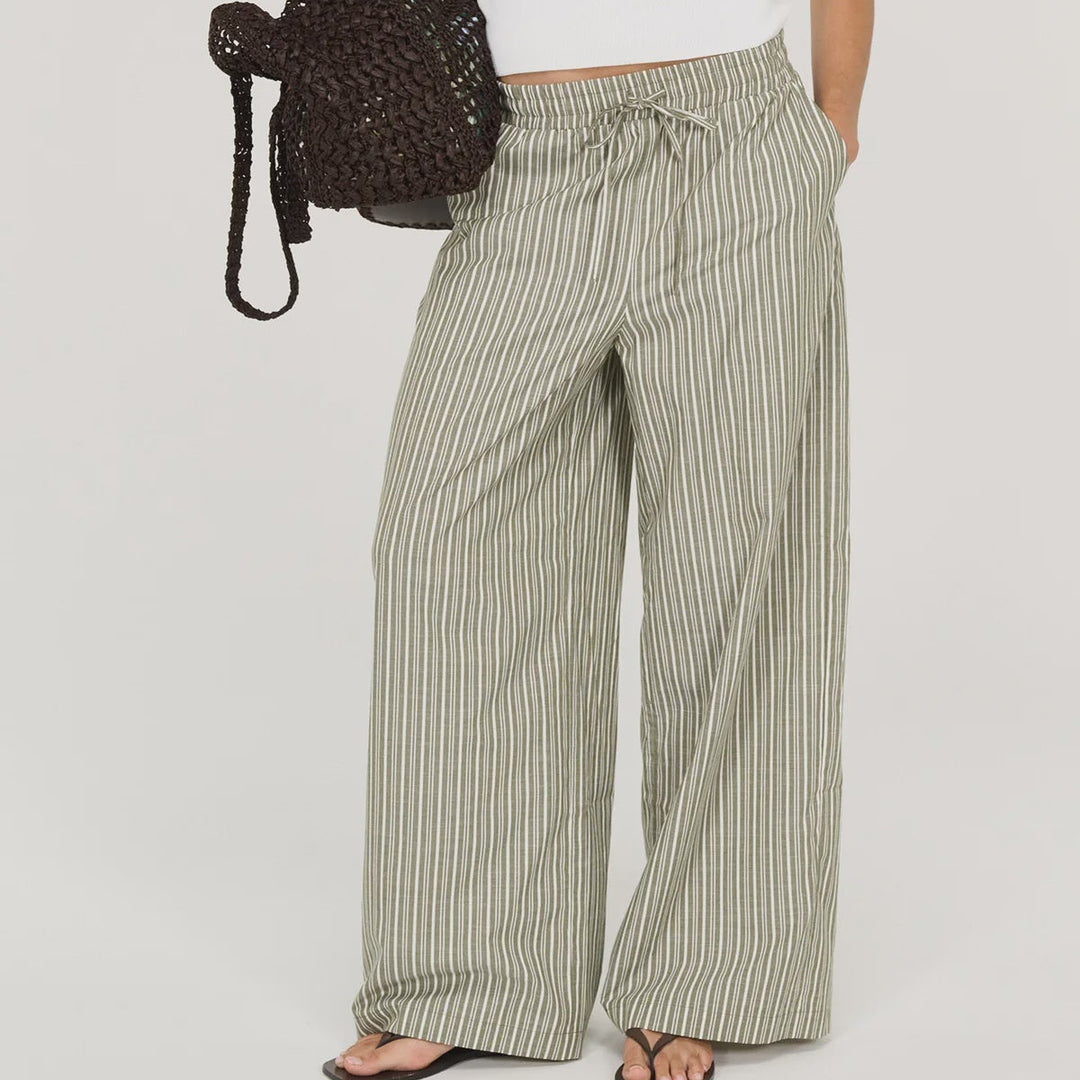 A woman modeling wide-leg striped pants in sage green and white, paired with a white sleeveless top, black sunglasses, and a dark woven handbag, against a plain white studio background.