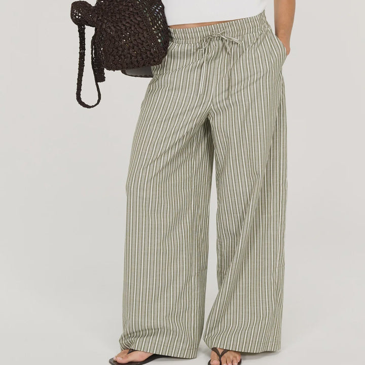 A woman modeling wide-leg striped pants in sage green and white, paired with a white sleeveless top, black sunglasses, and a dark woven handbag, against a plain white studio background.