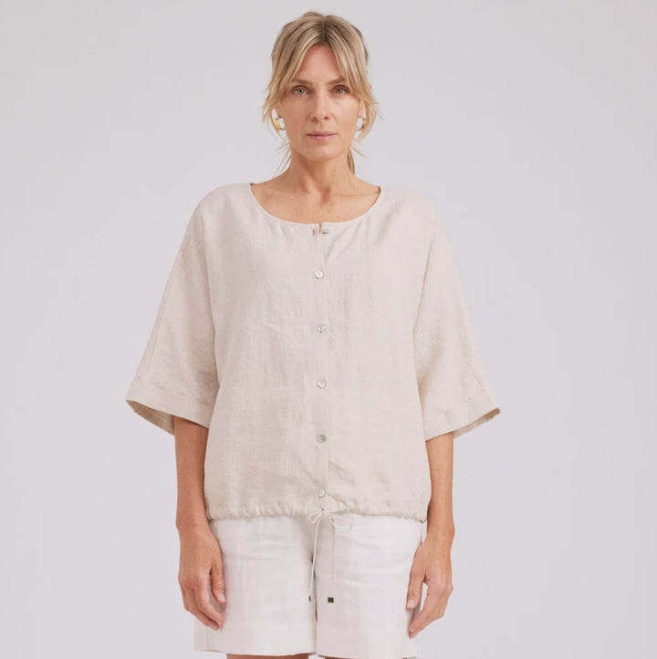 A woman modeling a light beige linen short-sleeve button-up top and matching off-white shorts against a plain light gray studio background.
