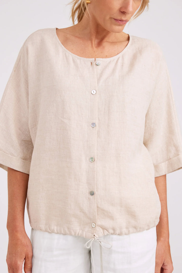 A woman wearing a light beige linen short-sleeve top with a button-up front and drawstring hem, paired with white pants, against a plain light background.