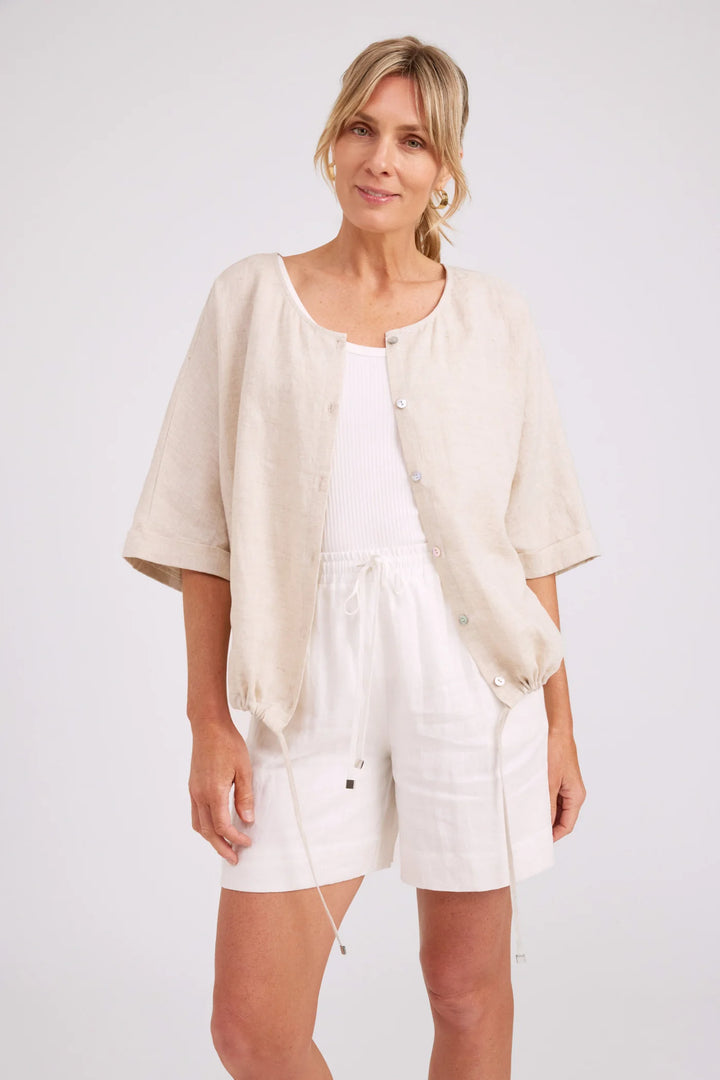 A woman modeling a light beige linen short-sleeve button-up cardigan over a white tank top and white shorts, photographed against a clean white studio background.
