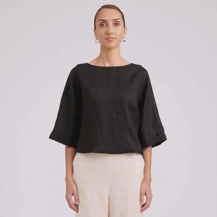 A model stands against a light gray studio background wearing a black short-sleeve top with wide draped sleeves and cream wide-leg trousers.