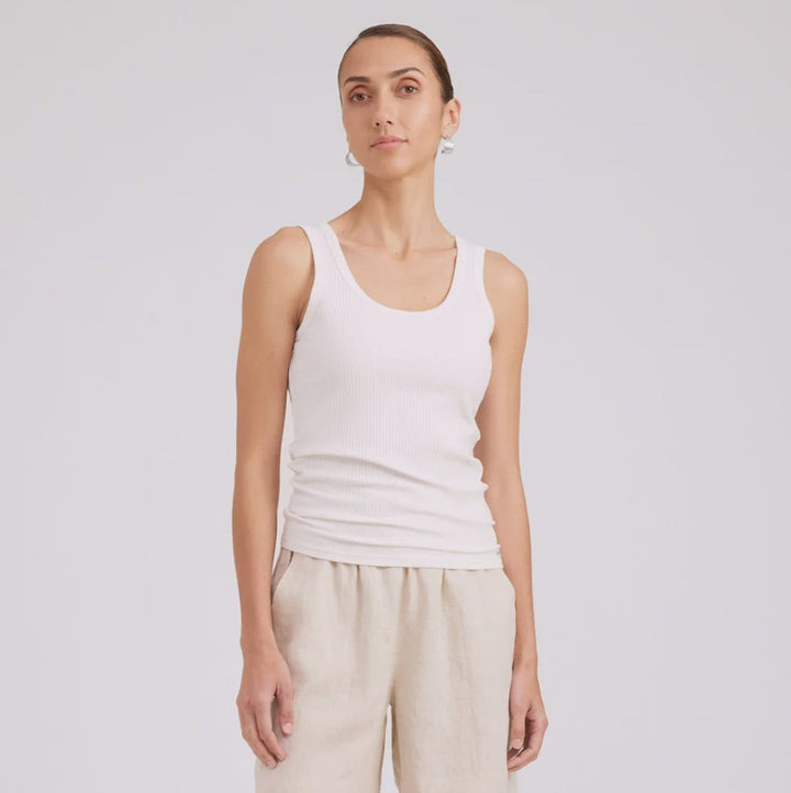 A woman modeling a white ribbed tank top and light beige linen shorts against a light gray studio background.
