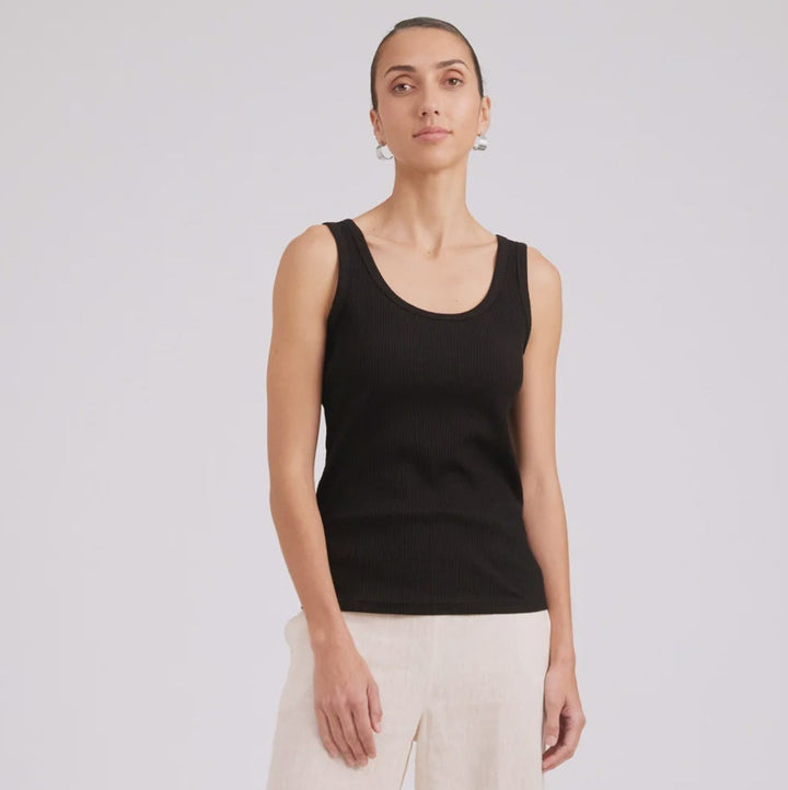 A woman modeling a black sleeveless tank top and wide-leg cream trousers with black sandals, standing against a light gray studio background.