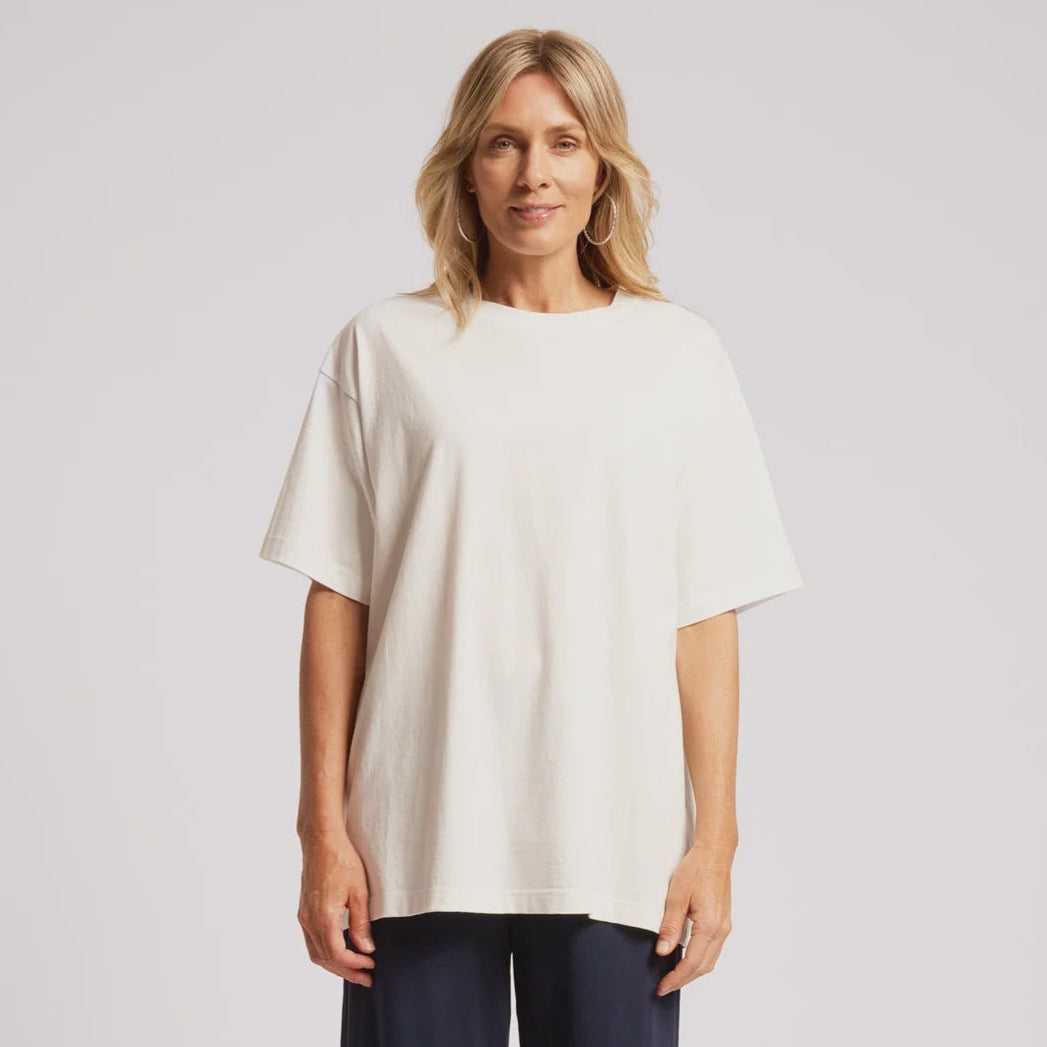 A woman modeling an oversized off-white short-sleeve top and dark navy wide-leg cropped pants with white sneakers, shot against a light gray studio background.