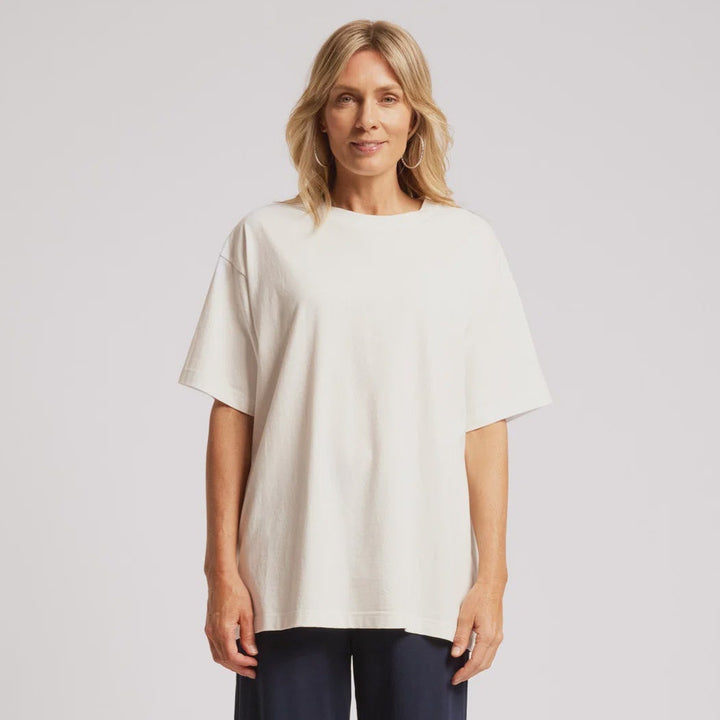 A woman modeling an oversized off-white short-sleeve top and dark navy wide-leg cropped pants with white sneakers, shot against a light gray studio background.