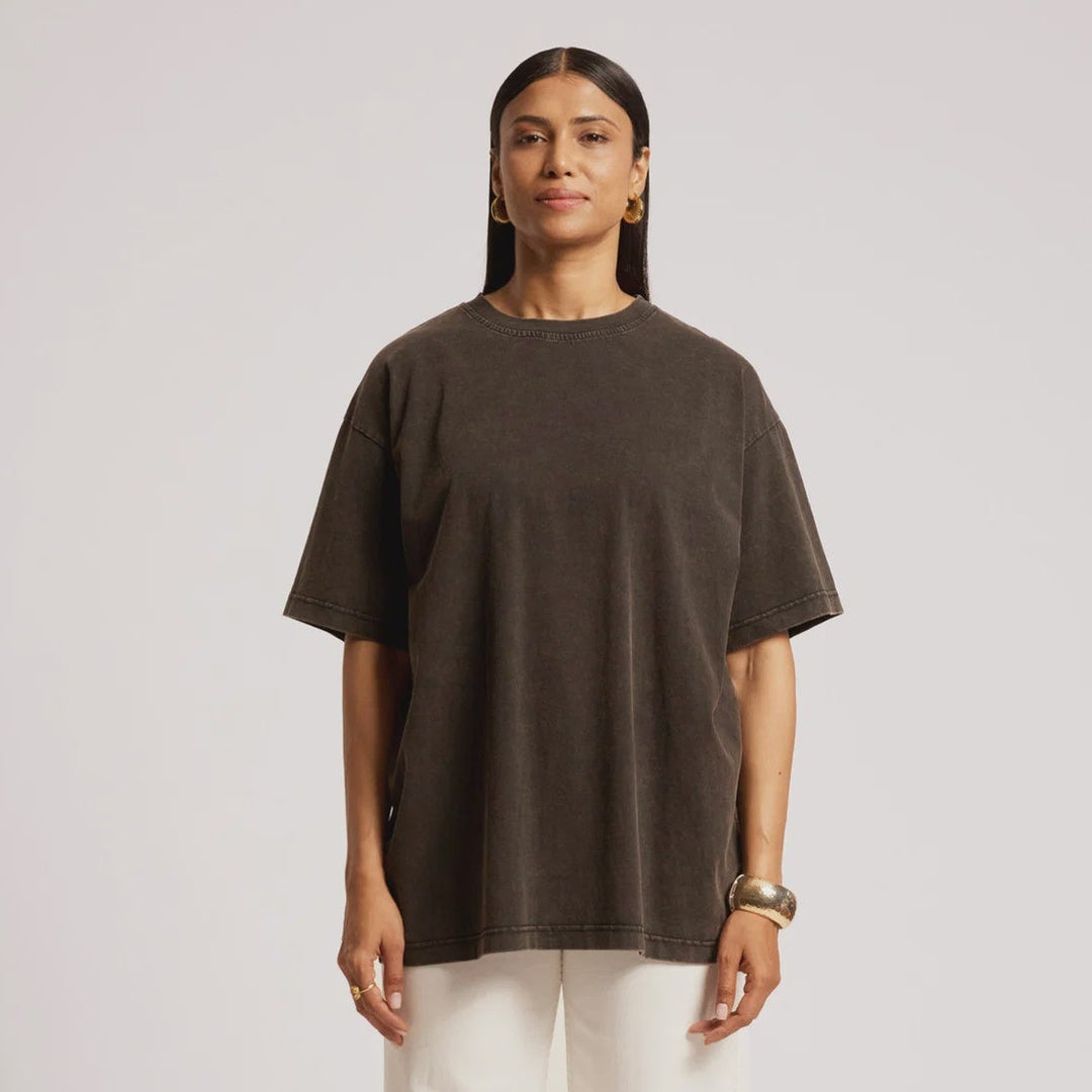 A woman stands against a light gray studio background wearing an oversized dark brown short-sleeve t-shirt, off-white wide-leg trousers, and brown textured shoes.