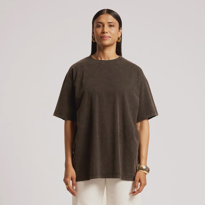 A woman stands against a light gray studio background wearing an oversized dark brown short-sleeve t-shirt, off-white wide-leg trousers, and brown textured shoes.