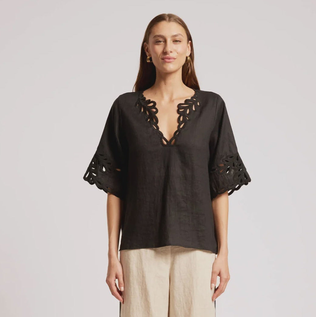 A woman modeling a black short-sleeve top with lace-up V-neck and floral lace sleeve cuffs, paired with wide-leg beige trousers and black sandals, against a light gray studio background.