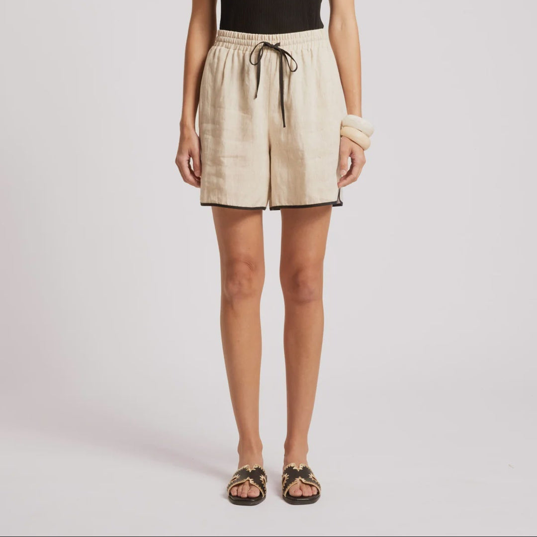 A model stands against a light gray studio background wearing a black sleeveless top and light beige drawstring shorts, with black sandals and a white wristband.