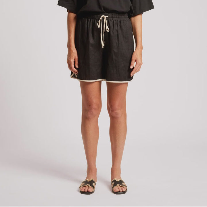 A woman modeling a black short-sleeve tunic top and matching black shorts with a cream drawstring, standing against a light gray studio background.