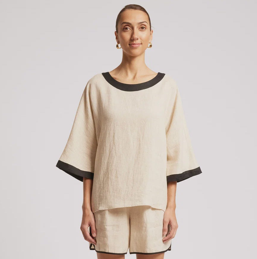 A woman modeling a matching beige linen lounge set with black trim, consisting of a short-sleeve top and shorts, against a plain light gray studio background.