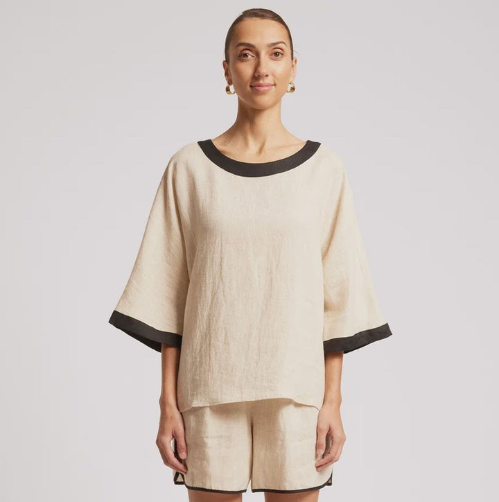 A woman modeling a matching beige linen lounge set with black trim, consisting of a short-sleeve top and shorts, against a plain light gray studio background.