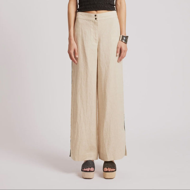 A woman stands against a light gray studio background wearing a black smocked sleeveless top and beige wide-leg trousers with side buttons and sandals.