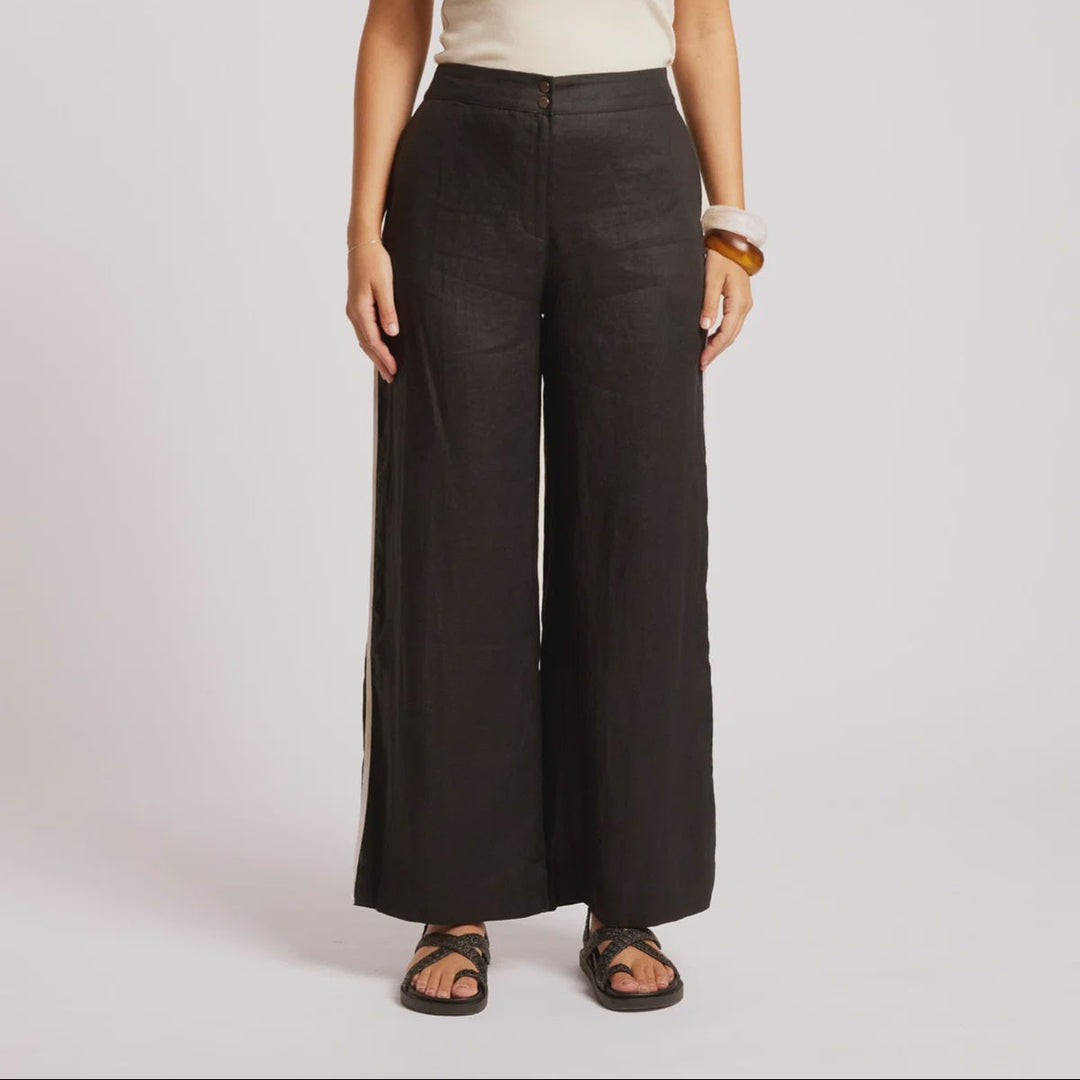 A woman stands against a light gray studio background wearing a cream ribbed tank top with black trim and black wide-leg trousers.