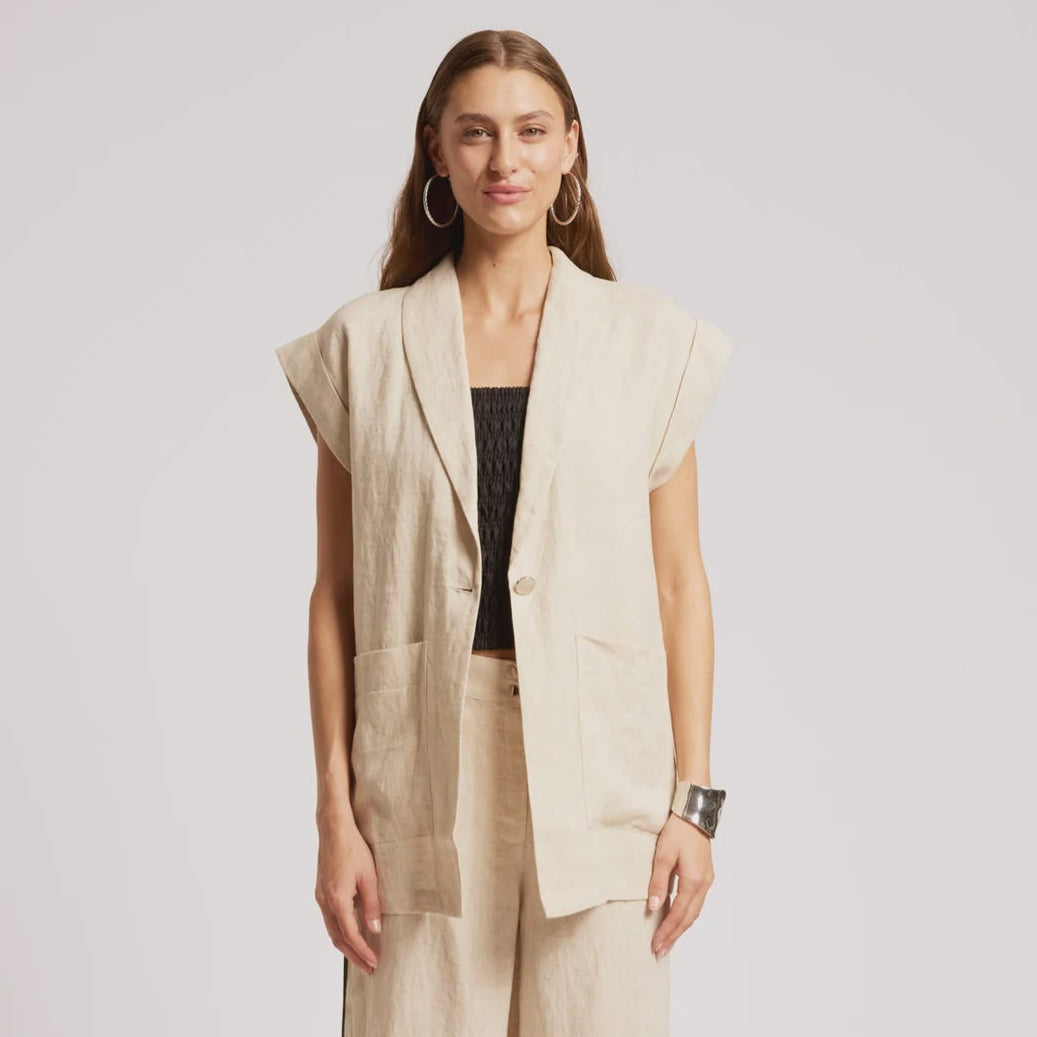 A woman modeling a light beige linen two-piece set consisting of a sleeveless button-front vest and matching wide-leg trousers, styled with a black ribbed top and black platform sandals against a light gray studio background.