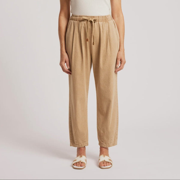 A woman modeling a pair of beige drawstring wide-leg pants with an elastic waist, styled with a white tank top and white slide sandals against a light gray studio background.