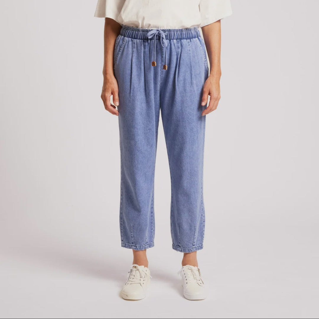 A woman modeling a pair of light blue drawstring cropped jeans with a white t-shirt and white sneakers, standing against a light gray studio background.