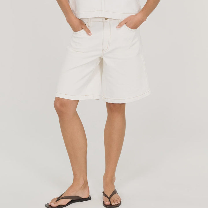 A woman modeling a white sleeveless halter crop top and matching white shorts, paired with black flip-flop sandals, against a light gray studio background.