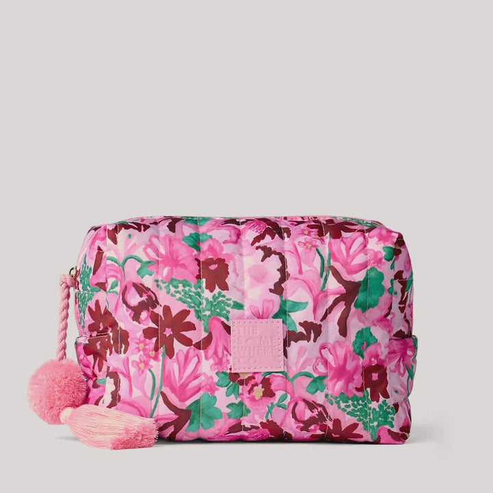 A pink nylon cosmetic pouch with a vibrant floral print in shades of pink, burgundy, and green, featuring a pink rope zipper pull adorned with two fluffy pink pom-poms.