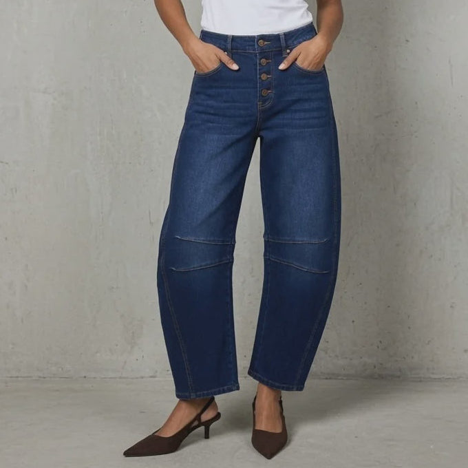 A woman stands against a textured light gray wall wearing dark blue wide-leg cropped jeans with a white sleeveless top and brown pointed-toe heels.