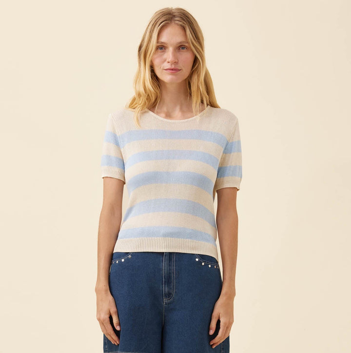 A woman stands against a plain warm beige studio background wearing a short-sleeve knit top with light blue and cream horizontal stripes, dark blue denim shorts, and tan slip-on shoes.