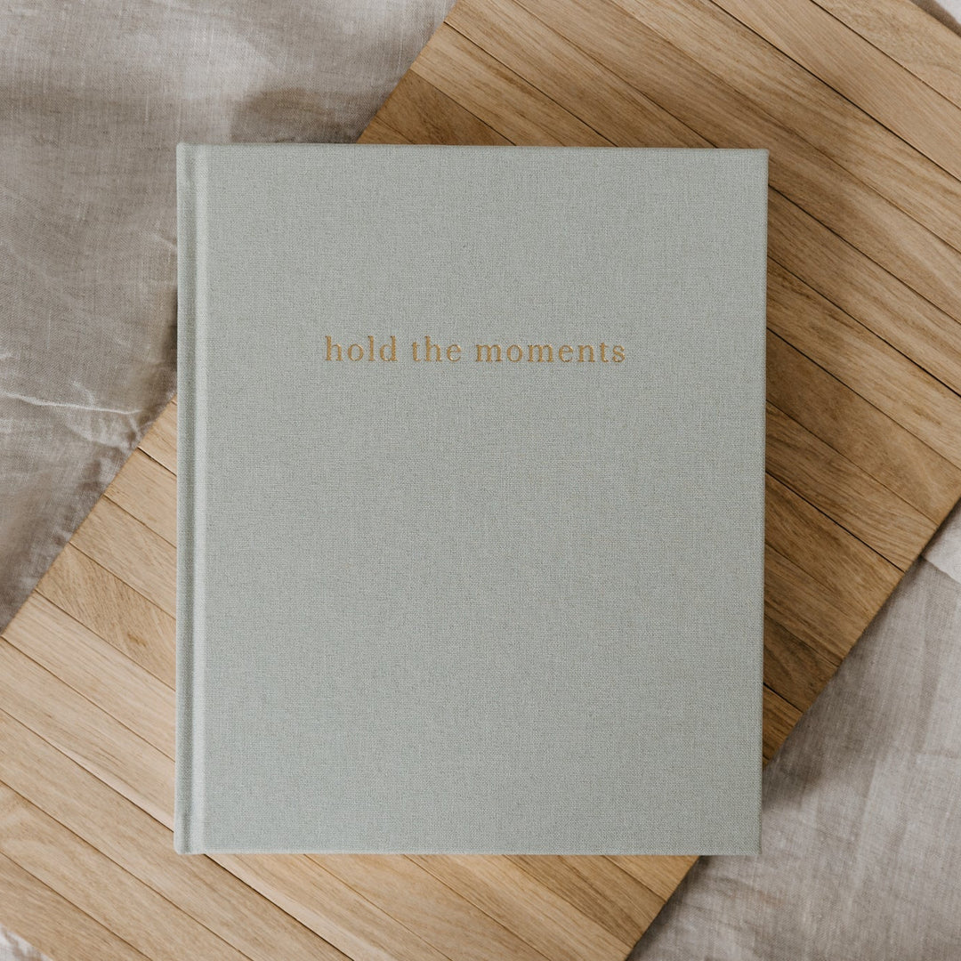 Light gray book titled 'hold the moments' on a wooden surface with soft fabric background
