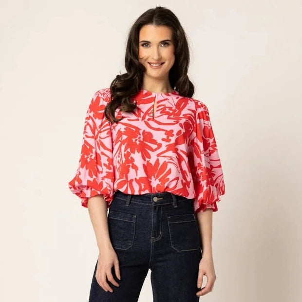 A woman modeling a red and white tropical floral print blouse with dark wash flare jeans and red sandals, standing against a plain beige studio background.