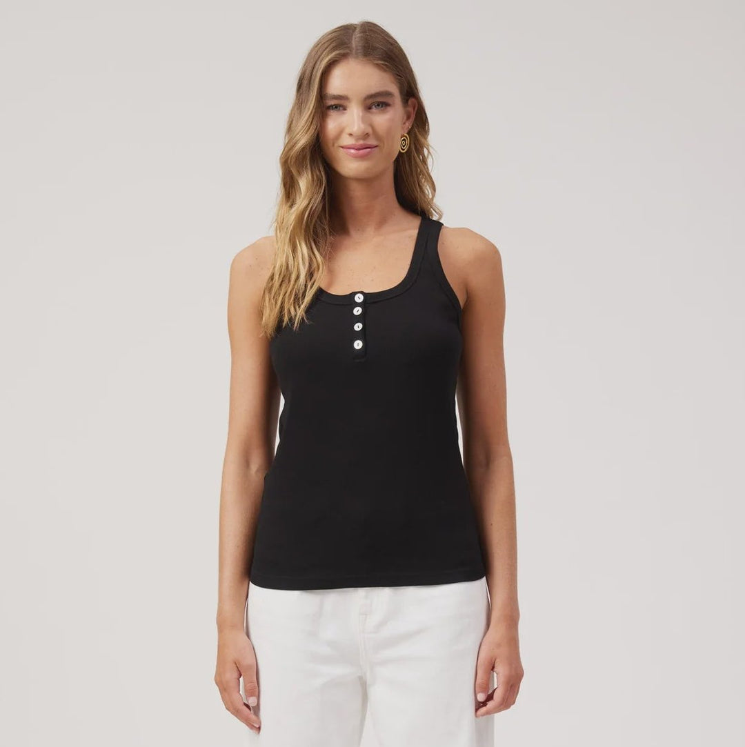A woman modeling a black sleeveless tank top with three white buttons at the neckline, paired with white wide-leg pants and brown sandals, against a plain white studio background.