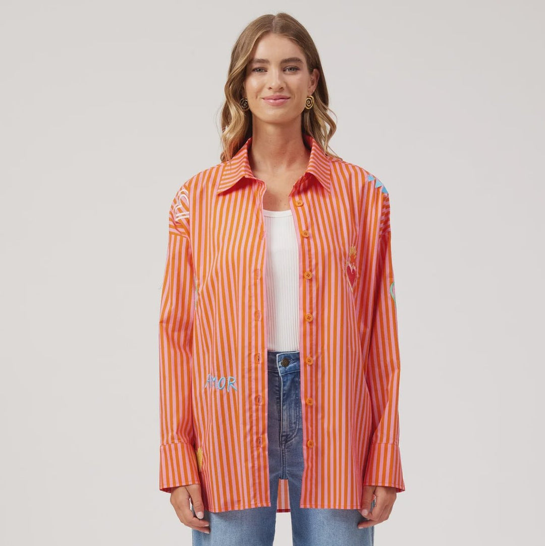 A woman stands against a light gray studio background wearing a bright orange and white vertically striped button-up shirt with embroidered details, a white top, wide-leg blue jeans, and tan sandals.