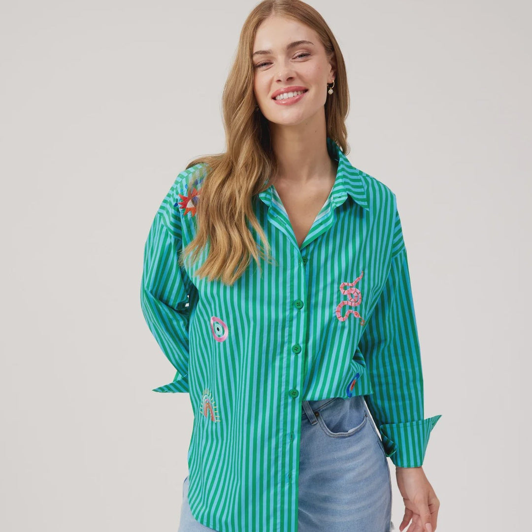 A woman modeling a green and white vertically striped button-up shirt with colorful floral embroidery on the front, paired with light-wash denim shorts, against a plain light gray studio background.