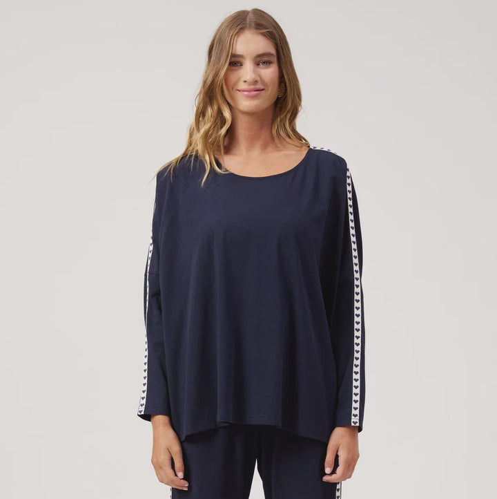 A woman modeling a matching navy blue lounge set consisting of a long-sleeve top with white patterned sleeve stripes and matching wide-leg pants with white patterned side stripes, set against a plain white studio background.