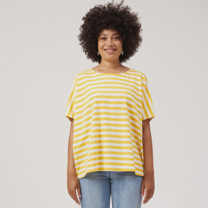A woman with an afro hairstyle stands against a light gray studio background wearing a yellow and white striped short-sleeve t-shirt, light blue jeans, and white sneakers.