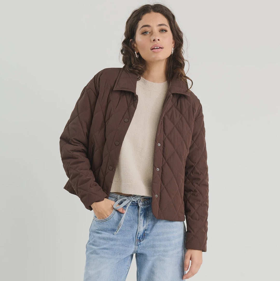 Woman wearing a brown quilted jacket, beige sweater, and light blue jeans on a white background