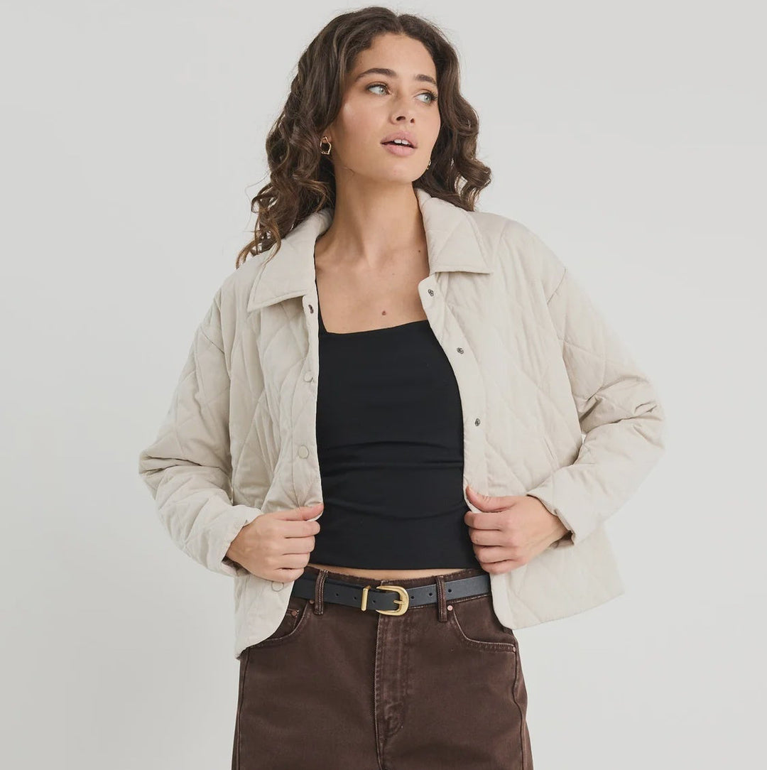Woman wearing a beige jacket, black top, and brown jeans on a white background