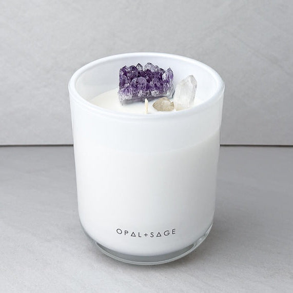 A white scented candle in a frosted glass jar with the brand name 'OPAL+SAGE' printed at the bottom. The candle is topped with a cluster of purple amethyst crystals and a clear quartz crystal, set against a neutral gray background.