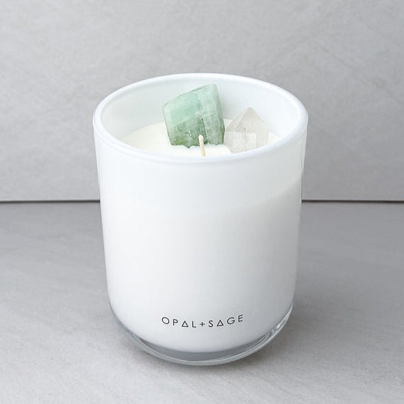 A white wax candle in a frosted glass jar with two crystal cubes inside, on a gray surface against a gray background. The brand name 'OPAL+SAGE' is printed on the front of the jar.