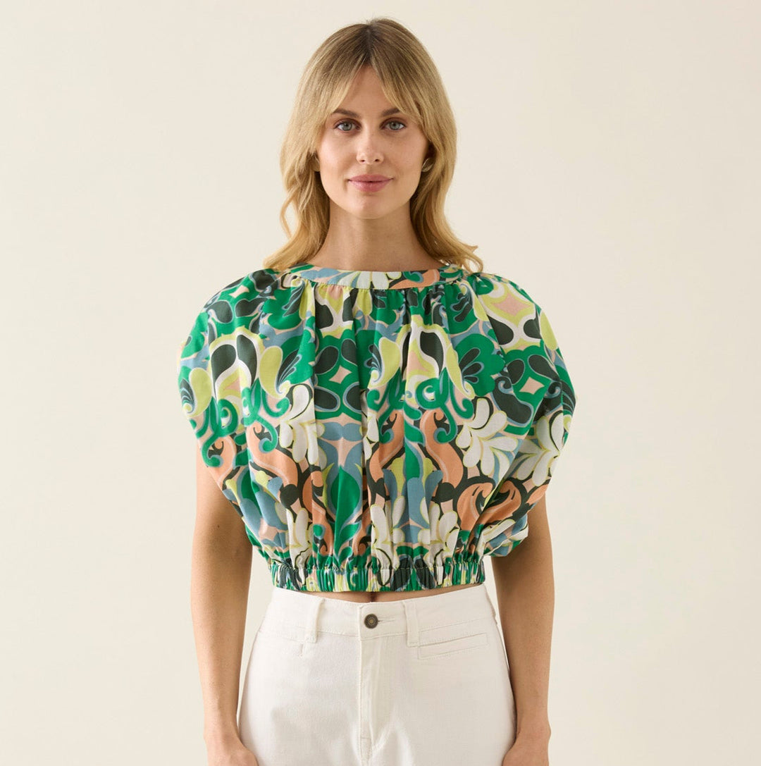 Woman wearing a patterned isle of mine top and white pants on a beige background