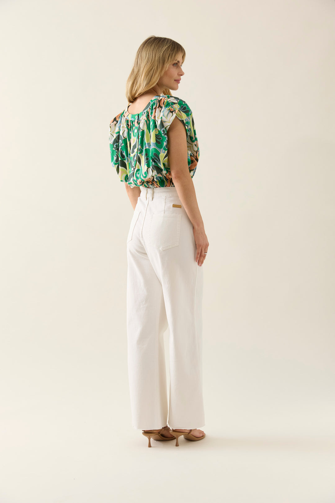Woman wearing a green isle of mine floral top and white pants on a plain background