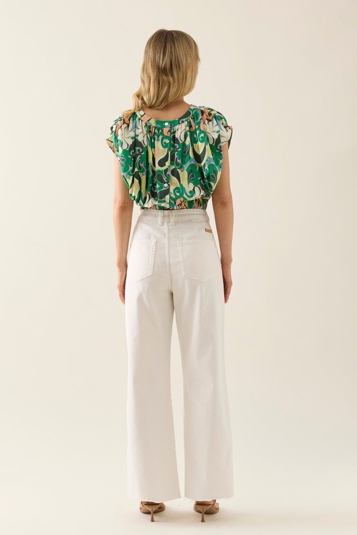 Woman wearing a patterned isle of mine top and white pants on a plain background
