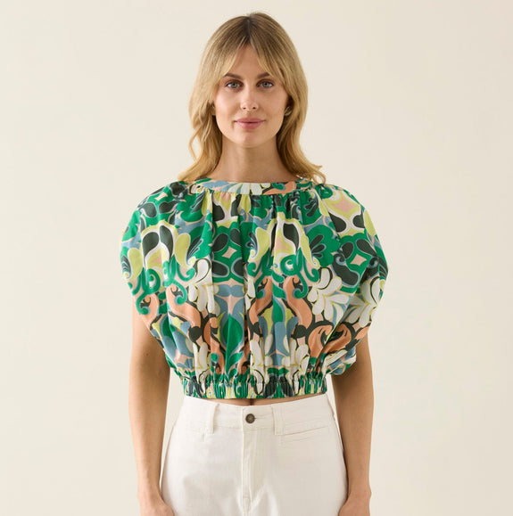 Woman wearing a patterned isle of mine top and white pants on a beige background