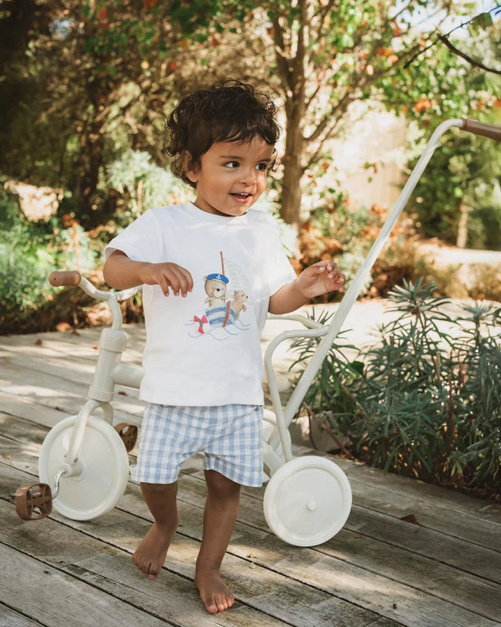 Child standing next to a white tricycle on a wooden deck with greenery in the background