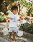 Child standing next to a white tricycle on a wooden deck with greenery in the background
