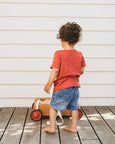 Child in a red  bebe shirt and blue shorts standing on a wooden deck with a toy.