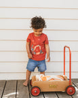 Child in a Bebe red shirt and denim shorts standing next to a brown wagon with wheels on a wooden deck.