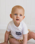 Baby wearing a white Bebe onesie with a raccoon design and brown shorts on a white background